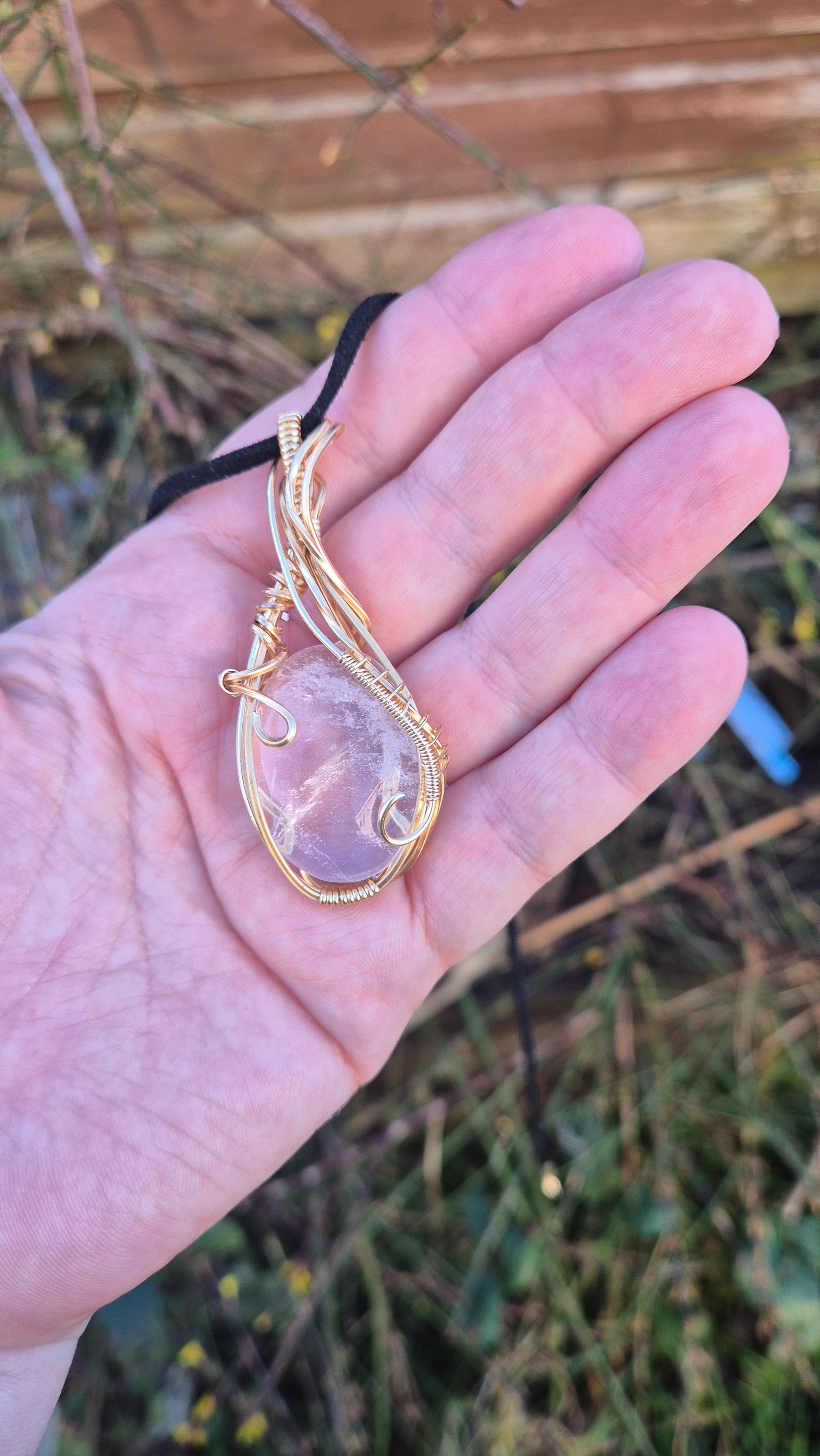 Hand holding a gold necklace with a clear quartz stone against a blurred natural background